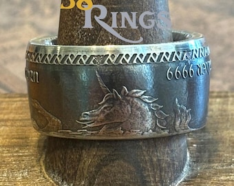 May include: A silver ring with a detailed unicorn design. The ring has a textured band with a repeating pattern and the text "6666 9999". The ring is displayed on a wooden stand.