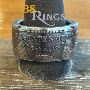 Op de afbeelding: Een zilveren ring gemaakt van een Amerikaanse dollarmunt. De ring heeft de woorden "United States of America" en "In God We Trust" op de band. De ring ligt op een houten oppervlak.
