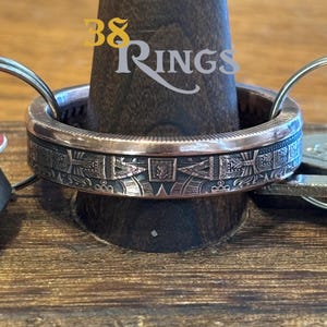 May include: A metallic ring featuring intricate engraved geometric patterns and a central square motif. The band has a copper-toned edge and a dark, oxidized finish in the recessed areas. The text 38 RINGS is overlaid on the image.