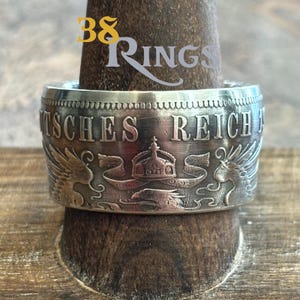 May include: A silver ring crafted from a vintage German coin. The coin features the inscription "Deutsches Reich" and a crown with two eagles flanking it.