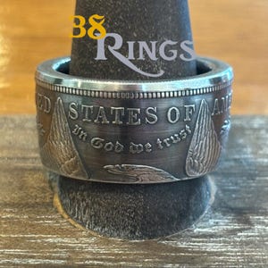 May include: A silver-toned metal ring crafted from a United States coin, featuring the embossed text "UNITED STATES OF AMERICA" and "IN GOD WE TRUST" along with detailed engravings of eagle wings and a bird head profile on the outer band surface.