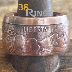 May include: A copper-colored ring with the word "LIBERTY" engraved on the side. The ring features a detailed mountain landscape design. The text "38 Rings" is visible at the top of the ring.