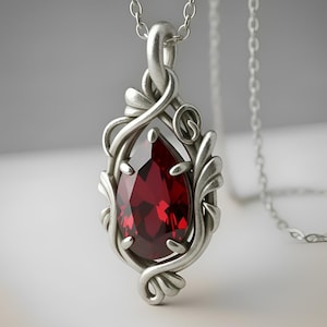May include: A silver pendant necklace featuring a teardrop-shaped, deep red gemstone. The gemstone is set within an ornate, swirling silver frame. The necklace hangs on a delicate silver chain, creating an elegant and eye-catching piece of jewelry.