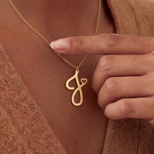 May include: A gold necklace featuring a cursive letter "J" pendant with a small heart. The chain is fine, and the pendant is the main focus. The necklace is worn by a person, highlighting a piece of personalized jewellery.