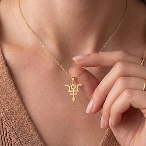 May include: A gold-toned necklace featuring a delicate chain and a pendant shaped like a stylized trident symbol with a circular top and a pointed base. The pendant hangs centrally from the chain, which rests against the skin of the neck.