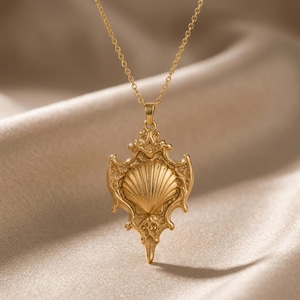 A highly detailed Sacred Shell Relic pendant, featuring a central ribbed scallop shell set within an elaborate, ornate frame with baroque-inspired scrollwork and pointed edges, hangs from a delicate cable chain. The pendant is shown in both rose gold and gold-plated finishes, resting on a light beige fabric surface next to dried floral elements, highlighting its intricate craftsmanship and vintage aesthetic.