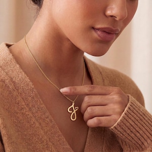 May include: A gold-toned necklace featuring a cursive letter J pendant with a small heart accent attached to the top right of the letter. The pendant hangs from a delicate gold-toned chain that rests against the collarbone area of the wearer.