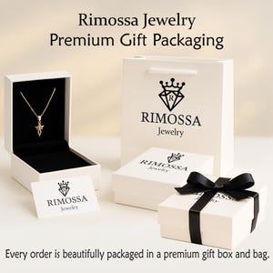 K&ouml;nnte beinhalten: Eine goldene Anh&auml;ngerkette wird in einer offenen wei&szlig;en Schmuckschatulle pr&auml;sentiert. Eine wei&szlig;e Geschenkt&uuml;te und zwei wei&szlig;e Schachteln mit dem Logo "Rimossa Jewelry" sind ebenfalls zu sehen. Eine Schachtel ist mit einem schwarzen Band verziert. Der Text lautet "Rimossa Jewelry Premium Gift Packaging."