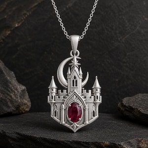 May include: A silver pendant necklace featuring a detailed castle structure with a crescent moon and star at the top. The center of the castle is adorned with a single, oval-shaped, deep red gemstone set in a polished silver frame.