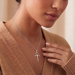 May include: A silver-toned necklace featuring a pendant shaped like an ankh symbol with a pointed, dagger-like base. The pendant hangs from a delicate, thin silver-toned chain that rests against the neck, showcasing a metallic finish.