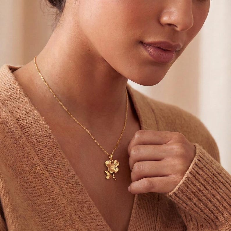 Pode incluir: Um colar dourado com um pingente de flor. O pingente apresenta um design floral detalhado com p&eacute;talas e um caule. O colar &eacute; usado por uma pessoa que veste um su&eacute;ter bege.