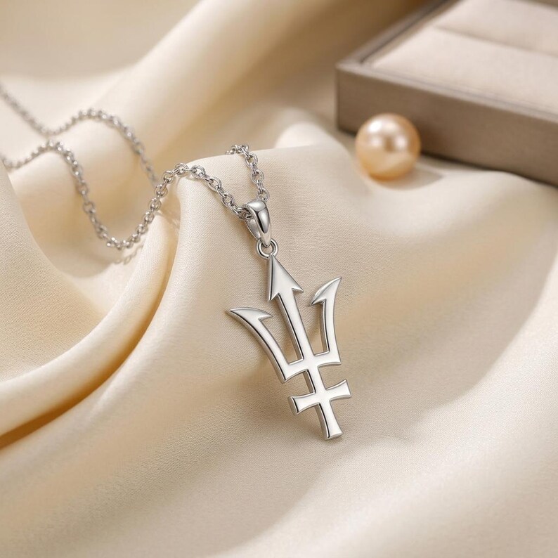 K&ouml;nnte beinhalten: Eine silberfarbene Halskette mit einem Dreizack-Anh&auml;nger. Der Anh&auml;nger hat ein dreizackiges Design mit einem Pfeil in der Mitte und einem Kreuz unten. Die Halskette wird auf cremefarbenem Satin pr&auml;sentiert.