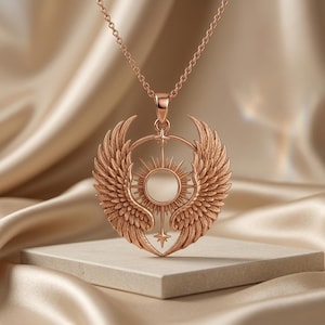 K&ouml;nnte beinhalten: Halskette mit Anh&auml;nger aus Ros&eacute;gold mit einem Sonnenmuster und detaillierten Fl&uuml;geln. Der Anh&auml;nger h&auml;ngt an einer zarten Kette. Die Halskette wird auf einer neutralen Oberfl&auml;che pr&auml;sentiert, um ihre komplizierten Details und ihren eleganten Stil hervorzuheben.