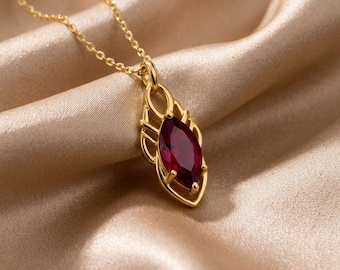 May include: A gold necklace featuring a marquise-cut deep red gemstone pendant. The pendant is held by a delicate gold chain and is encased in an intricate, open-work gold metal frame that follows the elongated shape of the central stone.