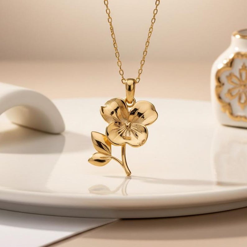 Pode incluir: Um colar com pingente dourado com um design floral detalhado. O pingente tem uma flor de tr&ecirc;s p&eacute;talas com duas folhas, suspensa por uma corrente delicada. O colar &eacute; exibido em uma superf&iacute;cie branca.