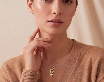 May include: A gold-toned necklace featuring a pendant with a cross symbol topped by a crescent moon shape. The pendant hangs from a delicate, thin gold-toned chain, resting against a light brown, textured knit sweater worn by an individual.