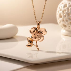 Pode incluir: Um colar com pingente em ouro rosa com um design de flor e folha. O pingente de flor tem p&eacute;talas detalhadas e &eacute; suspenso por uma corrente delicada. O colar &eacute; exibido em uma superf&iacute;cie branca.