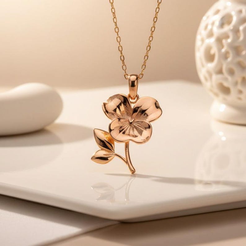 Pode incluir: Um colar com pingente em ouro rosa com um design de flor e folha. O pingente de flor tem p&eacute;talas detalhadas e &eacute; suspenso por uma corrente delicada. O colar &eacute; exibido em uma superf&iacute;cie branca.