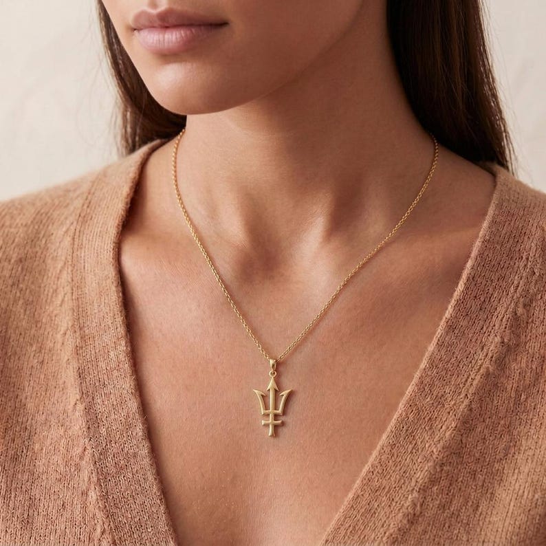 May include: A gold-colored necklace with a trident pendant. The pendant is a stylized trident design, suspended from a delicate chain. The necklace is worn against a neutral-toned background.