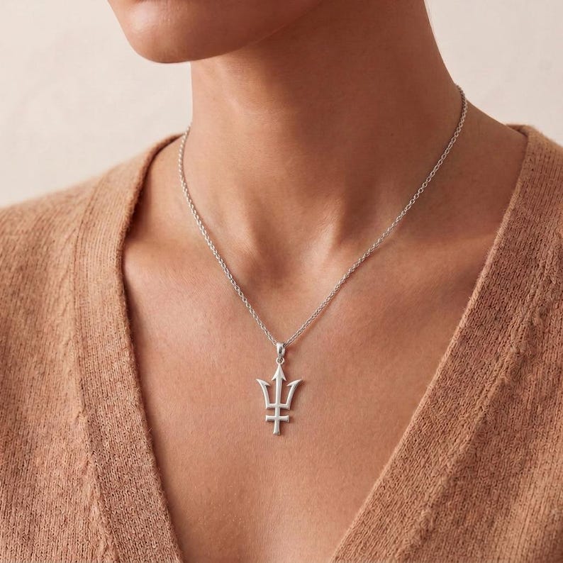 K&ouml;nnte beinhalten: Eine silberfarbene Halskette mit einem Dreizack-Anh&auml;nger. Der Anh&auml;nger h&auml;ngt an einer feinen Kette. Die Halskette wird auf der Haut getragen. Der Schmuck ist ein schlichtes, elegantes St&uuml;ck.
