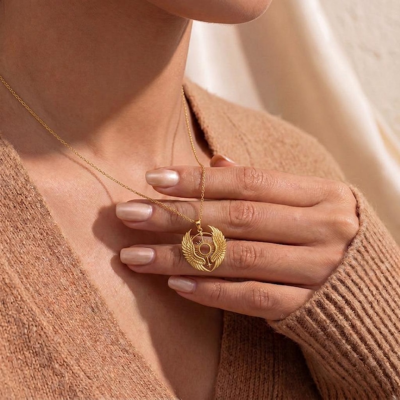 K&ouml;nnte beinhalten: Eine goldfarbene Halskette mit einem Anh&auml;nger im &auml;gyptischen Stil. Der Anh&auml;nger zeigt eine Sonnenscheibe mit Fl&uuml;geln und anderen Symbolen. Die Halskette wird von einer Person mit einem beigefarbenen Pullover getragen.