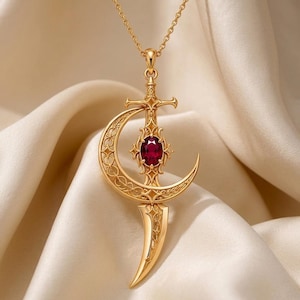 May include: A gold-toned pendant necklace featuring a sword design integrated with a crescent moon. The sword hilt is adorned with an oval-shaped, deep red gemstone at its center. The metalwork displays intricate filigree patterns throughout.
