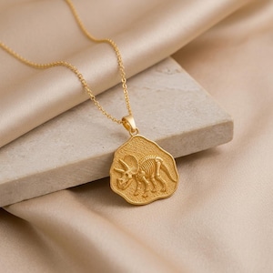May include: A gold-toned necklace featuring a pendant with an irregular, organic shape. The pendant displays a detailed, embossed skeleton of a triceratops dinosaur on a textured surface, suspended from a delicate, matching gold-toned chain.