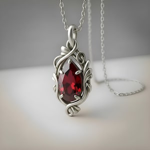 May include: A silver pendant necklace featuring a pear-shaped, deep red gemstone set within an intricate, swirling metal frame. The pendant hangs from a delicate silver chain, showcasing a polished finish and detailed metalwork surrounding the stone.