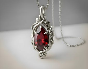 Puede incluir: Un collar con colgante de plata que presenta una piedra preciosa de color rojo intenso en forma de pera, engastada en un marco de metal con motivos de remolinos. El colgante cuelga de una fina cadena de plata, con un acabado pulido y detallado.