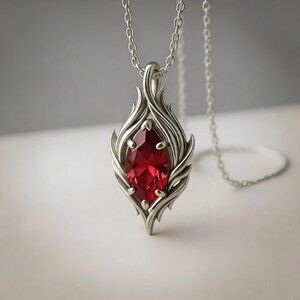 May include: A silver-toned pendant necklace featuring a marquise-cut deep red gemstone held by four prongs. The metal setting has a flame-like or leaf-like design with curved, flowing lines that frame the central stone. A delicate chain is attached.