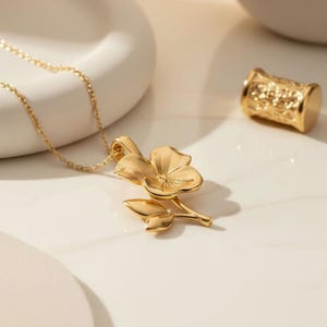 Pode incluir: Um colar dourado com um pingente floral. O pingente apresenta um design floral detalhado com p&eacute;talas e folhas. Um pequeno cilindro dourado texturizado tamb&eacute;m &eacute; vis&iacute;vel.