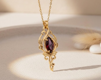 May include: A gold-toned necklace featuring a marquise-cut deep red gemstone set within an intricate, openwork metal frame. The pendant is suspended from a delicate gold-toned chain, showcasing a detailed, swirling filigree design.