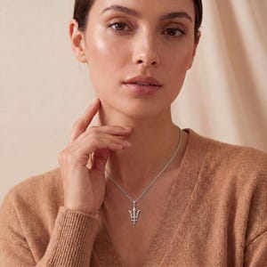 K&ouml;nnte beinhalten: Eine silberne Halskette mit einem Anh&auml;nger in Form eines Dreizacks. Der Anh&auml;nger h&auml;ngt an einer feinen Kette und befindet sich mittig auf der Brust. Die Frau tr&auml;gt einen braunen Pullover.