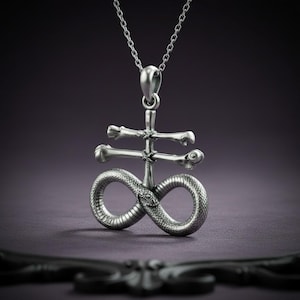 May include: A silver-toned pendant necklace featuring a vertical central bar crossed by two horizontal bone-shaped bars, with a snake coiled at the base into an infinity symbol shape. The pendant hangs from a delicate silver-toned chain link necklace.