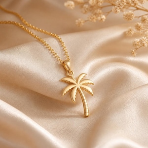 May include: A gold-toned necklace featuring a palm tree pendant. The pendant is shaped like a palm tree with detailed fronds and a textured trunk. The necklace consists of a delicate gold-toned chain that connects to the top of the pendant.