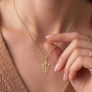 K&ouml;nnte beinhalten: Eine goldfarbene Halskette mit Anh&auml;nger in Form eines Dreizacks. Der Anh&auml;nger h&auml;ngt an einer filigranen Kette. Der Dreizack hat drei Zinken oben und einen horizontalen Balken unten. Die Halskette wird vor einem neutralen Hintergrund getragen.