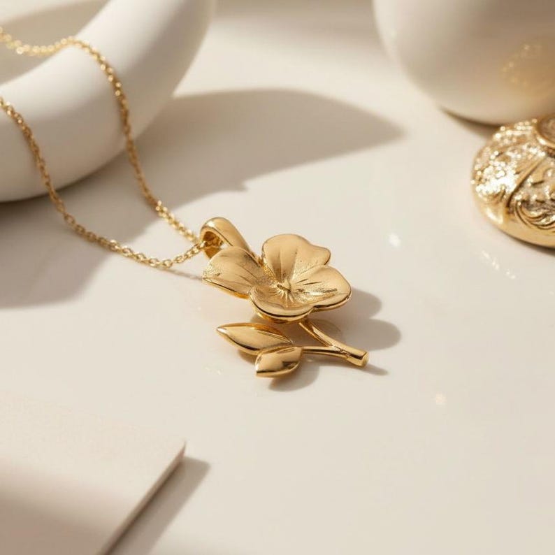 Pode incluir: Um colar dourado com um pingente de flor. O pingente apresenta um design floral detalhado com p&eacute;talas e folhas. A corrente &eacute; delicada e o pingente &eacute; centralizado, criando um visual elegante.