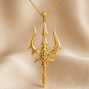 May include: A gold-toned pendant shaped like a three-pronged trident, featuring intricate filigree and scrollwork detailing throughout the metal surface. The pendant is suspended from a matching gold-toned chain, showcasing a polished finish.
