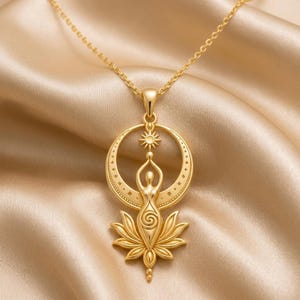May include: A gold-toned necklace featuring a pendant with a central figure standing above a lotus flower, positioned within a crescent moon frame. The figure has a spiral design on the torso, and a small sun symbol sits above the head, all suspended from a delicate gold chain.