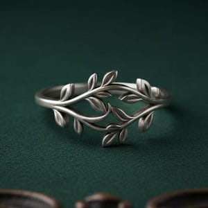 May include: A silver-toned ring featuring a band designed with a series of interconnected, sculpted leaves that wrap around the finger. The metal has a matte, slightly oxidized finish that highlights the detailed veins and shapes of the foliage.