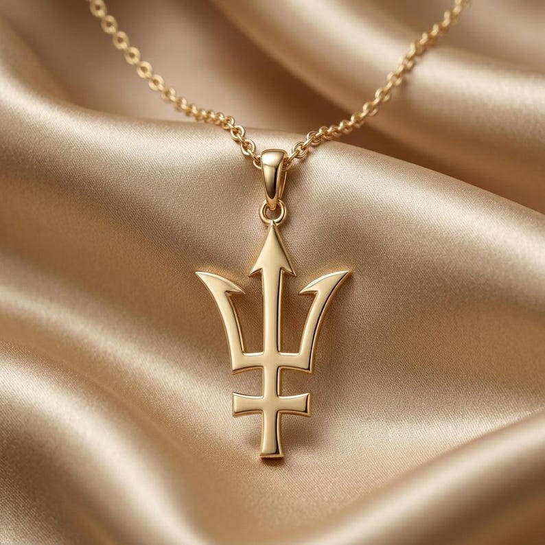 K&ouml;nnte beinhalten: Eine goldfarbene Halskette mit Anh&auml;nger, die ein Dreizack-Design aufweist. Der Anh&auml;nger h&auml;ngt an einer zarten Kette vor einem Hintergrund aus drapiertem, goldenem Stoff. Der Dreizack hat einen zentralen pfeilf&ouml;rmigen Zacken und zwei gebogene Seitenzacken.