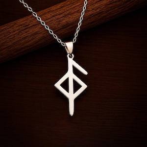 Könnte beinhalten: Silberfarbene Anhängerkette mit einem stilisierten Runensymbol. Der Anhänger hängt an einer filigranen Kette und ist vor einem dunkelbraunen Holzgrund platziert. Die Rune ist ein geometrisches Design mit einer Raute und einer vertikalen Linie.