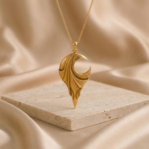 May include: A gold-toned pendant necklace featuring a crescent moon shape with textured, wing-like drapery extending downward to a point. The pendant hangs from a delicate gold-toned chain, showcasing a metallic finish and intricate sculptural details.