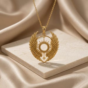 K&ouml;nnte beinhalten: Goldfarbene Anh&auml;ngerkette mit detailliertem Sonnenmuster und stilisierten Fl&uuml;geln. Der Anh&auml;nger h&auml;ngt an einer filigranen Kette. Die Halskette wird auf einer neutralen Oberfl&auml;che pr&auml;sentiert.
