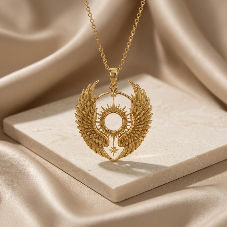 K&ouml;nnte beinhalten: Goldfarbene Anh&auml;ngerkette mit detailliertem Sonnenmuster und stilisierten Fl&uuml;geln. Der Anh&auml;nger h&auml;ngt an einer filigranen Kette. Die Halskette wird auf einer neutralen Oberfl&auml;che pr&auml;sentiert.