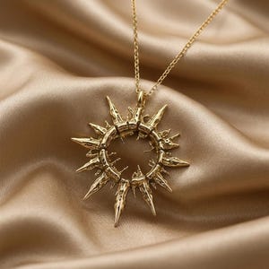 May include: A gold-toned necklace featuring a circular pendant with a sunburst design. The pendant has a textured, irregular surface with multiple pointed rays extending outward from the center ring, suspended from a delicate gold-toned chain.