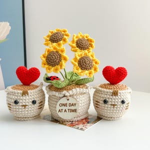 以下が含まれることがあります： 黄色の花びらと茶色の中心部を持つかぎ針編みのヒマワリのアレンジメント。白い鉢植えには「ONE DAY AT A TIME」と書かれた木のタグが付いています。赤いハートのトッパーが付いた2つの小さなマッチングポットがアレンジメントを挟んでいます。ポットには猫の顔のデザインがあります。