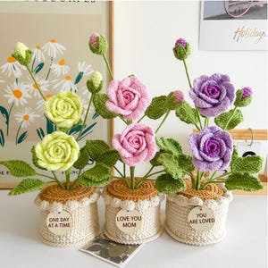 Puede incluir: Tres arreglos florales de ganchillo en macetas color crema. Cada maceta tiene un mensaje diferente en una etiqueta de madera: "ONE DAY AT A TIME", "LOVE YOU MOM" y "YOU ARE LOVED". Las flores son en tonos verdes, rosas y morados.