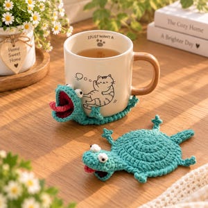 Peut inclure: Un mug en céramique avec une anse marron et un dessin au trait noir représentant un chat, avec le texte I JUST WANT A et une empreinte de patte. Le mug est entouré d'une housse en crochet bleu sarcelle en forme de grenouille avec une langue rouge.
