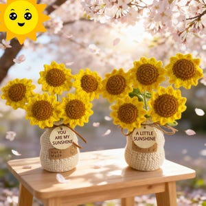 以下が含まれることがあります： 小さなジュートの花瓶に入った、手作りのかぎ針編みのヒマワリのブーケが2つ。ヒマワリは黄色い花びらと茶色の中心部をしています。1つの花瓶には「YOU ARE MY SUNSHINE」、もう1つには「A LITTLE SUNSHINE」と書かれたタグが付いています。